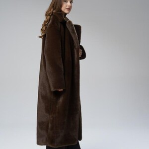 May include: A long, brown faux fur coat with a wide collar. The coat is floor-length and worn over black pants and shoes. The model is facing the camera, with a neutral background.
