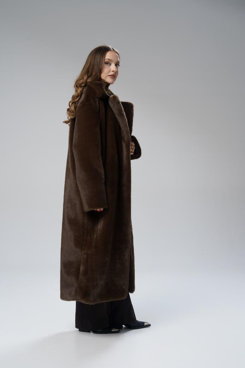 May include: A long, brown faux fur coat with a wide collar. The coat is floor-length and worn over black pants and shoes. The model is facing the camera, with a neutral background.