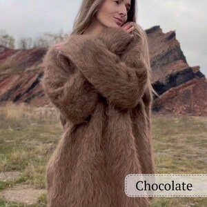 Puede incluir: Un su&eacute;ter largo de color chocolate con una textura esponjosa. El su&eacute;ter lo lleva una persona. La palabra "Chocolate" se muestra en un rect&aacute;ngulo blanco en la parte inferior derecha de la imagen. El fondo presenta un entorno natural al aire libre.