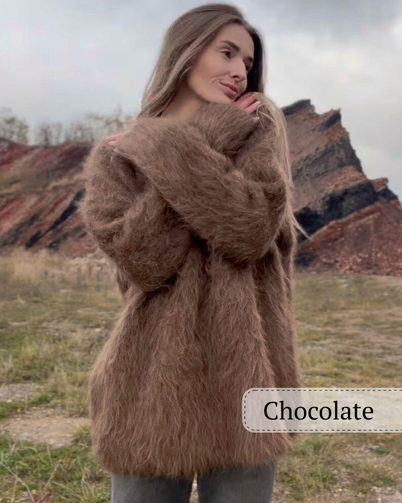 Puede incluir: Un su&eacute;ter largo de color chocolate con una textura esponjosa. El su&eacute;ter lo lleva una persona. La palabra "Chocolate" se muestra en un rect&aacute;ngulo blanco en la parte inferior derecha de la imagen. El fondo presenta un entorno natural al aire libre.