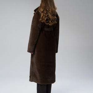 May include: A long, dark brown coat with a belt, worn over dark pants. The coat has a wide collar and long sleeves. The model is facing away from the camera, with long, wavy brown hair.
