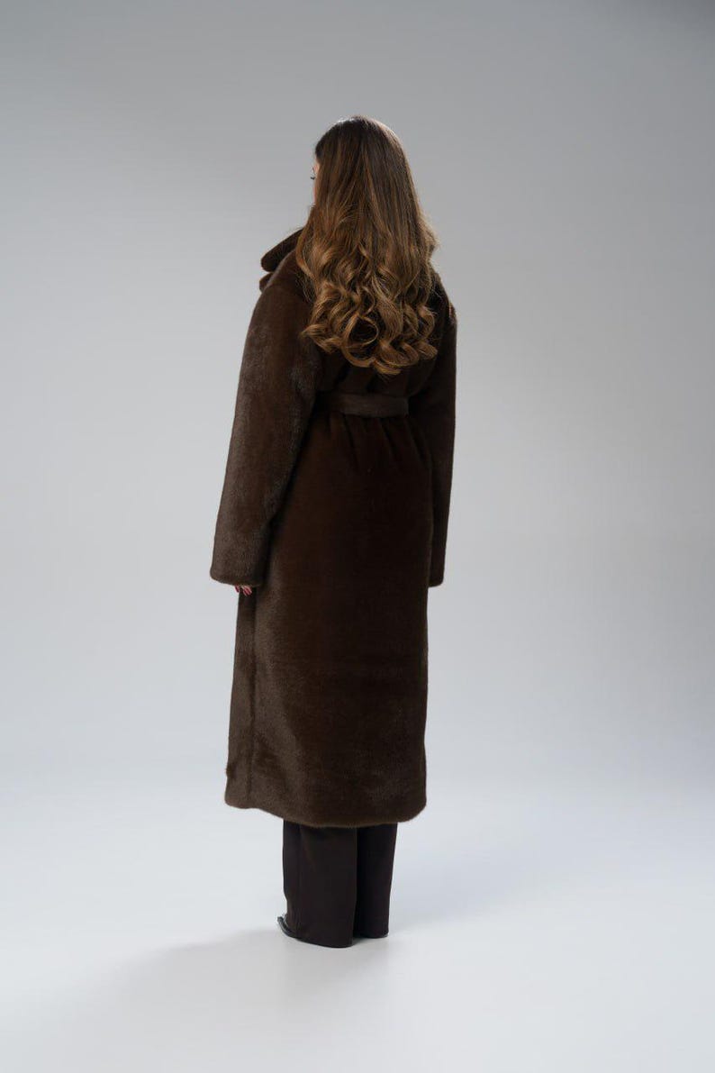 May include: A long, dark brown coat with a belt, worn over dark pants. The coat has a wide collar and long sleeves. The model is facing away from the camera, with long, wavy brown hair.