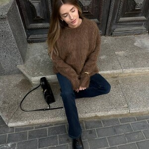 Puede incluir: Un su&eacute;ter marr&oacute;n y difuso combinado con vaqueros azul oscuro y botas negras. Un bolso negro con una correa larga descansa sobre los escalones. La persona est&aacute; sentada en los escalones frente a una puerta de madera oscura.