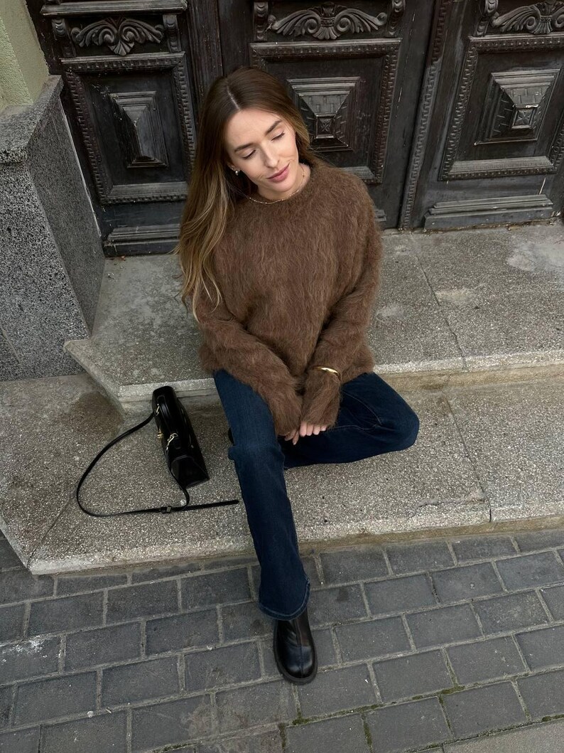 Puede incluir: Un su&eacute;ter marr&oacute;n y difuso combinado con vaqueros azul oscuro y botas negras. Un bolso negro con una correa larga descansa sobre los escalones. La persona est&aacute; sentada en los escalones frente a una puerta de madera oscura.