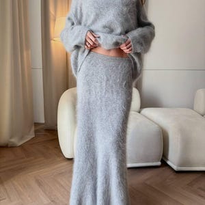 May include: A light gray, fuzzy knit sweater and matching maxi skirt set. The sweater has a crew neck and long sleeves. The skirt is a high-waisted, floor-length design. The outfit is made of a soft, textured material, perfect for a cozy, stylish look.