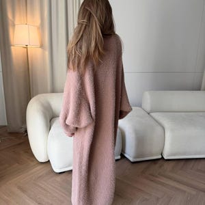 May include: A long, dusky pink cardigan with a soft, textured appearance. The cardigan has long sleeves and a floor-length design. The person is facing away from the camera, showcasing the garment's drape and style. The cardigan is a cosy and stylish outerwear option.