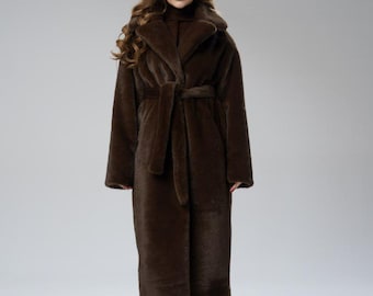 May include: A long, brown faux fur coat with a shawl collar and a matching belt. The coat is floor-length and has a soft, textured appearance. The model is wearing black trousers and a brown roll neck.