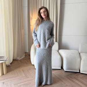 Peut inclure: Ensemble assorti, pull et jupe gris clair. Le pull à manches longues a un col rond et la jupe est longue jusqu'au sol. La tenue est faite d'une matière douce et duveteuse. Le fond comprend un canapé blanc et des rideaux clairs.