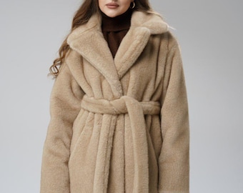 May include: A long, beige faux fur coat with a shawl collar and a matching belt. The coat is worn over a brown roll neck. The coat has a soft, plush texture and a classic, elegant design.