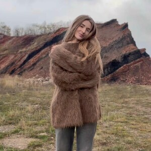 Puede incluir: Una persona con un su&eacute;ter marr&oacute;n y difuso y unos vaqueros grises. El su&eacute;ter tiene un corte largo y holgado. El fondo presenta un cielo nublado y una ladera rocosa de color marr&oacute;n rojizo.