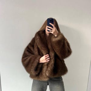 May include: A brown faux fur coat with a wide collar and long sleeves. The coat is a rich, warm brown color and appears soft and plush. The person is wearing gray jeans.