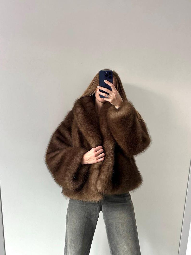 May include: A brown faux fur coat with a wide collar and long sleeves. The coat is a rich, warm brown color and appears soft and plush. The person is wearing gray jeans.