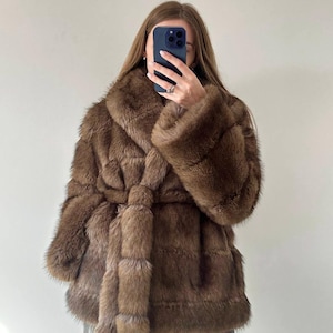 May include: A brown faux fur coat with a wide collar and a matching belt tied at the waist. The coat has long sleeves and a textured appearance. The coat is a medium brown color.