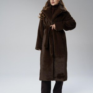 May include: A long, dark brown faux fur coat with a matching belt. The coat has a wide collar and long sleeves. The model is wearing dark brown pants and black shoes. The coat is a full-length design.