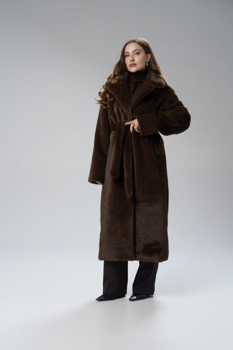 May include: A long, dark brown faux fur coat with a matching belt. The coat has a wide collar and long sleeves. The model is wearing dark brown pants and black shoes. The coat is a full-length design.