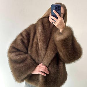 May include: A brown faux fur coat with a wide collar and long sleeves. The coat has a soft, plush texture and a relaxed fit. The coat is a medium brown color.