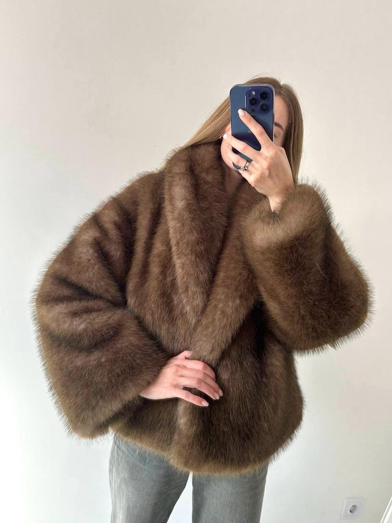 May include: A brown faux fur coat with a wide collar and long sleeves. The coat has a soft, plush texture and a relaxed fit. The coat is a medium brown color.