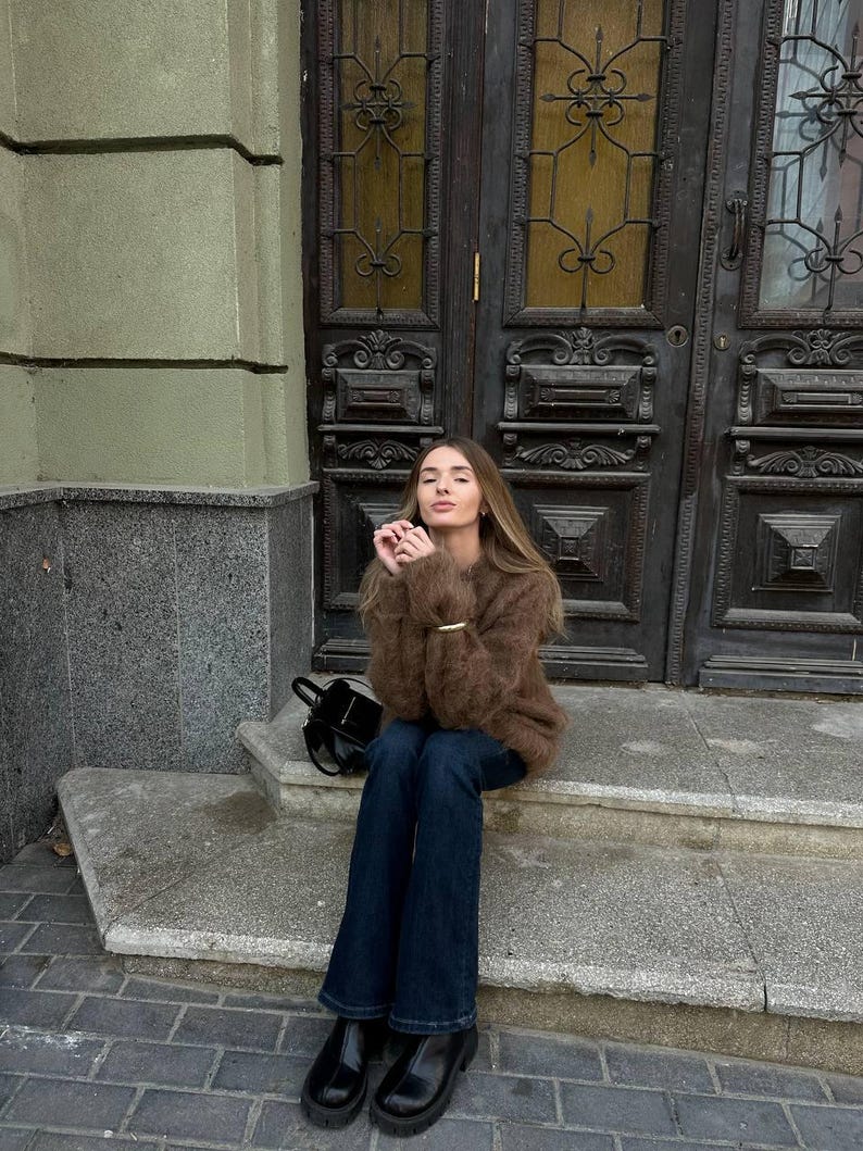 Puede incluir: Una persona con un su&eacute;ter marr&oacute;n difuso, jeans azul oscuro y botas negras, sentada en escalones de piedra. Un peque&ntilde;o bolso negro descansa cerca. El fondo presenta una puerta de madera oscura y una pared de piedra gris.