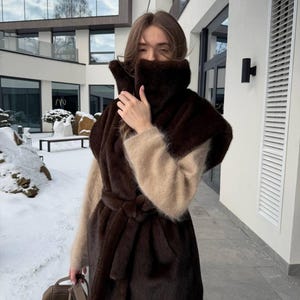 Könnte beinhalten: Eine Frau trägt eine braune Kunstfellweste mit passendem Gürtel und einen beigen Pullover. Die Weste hat einen hohen Kragen und wird mit einer weißen Hose und einer braunen Handtasche kombiniert. Der Hintergrund zeigt eine verschneite Umgebung im Freien.