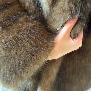 May include: Close-up of a brown fur coat. The coat has a soft, textured appearance with varying shades of brown. A hand is holding the coat, and a small, rectangular tag is visible.