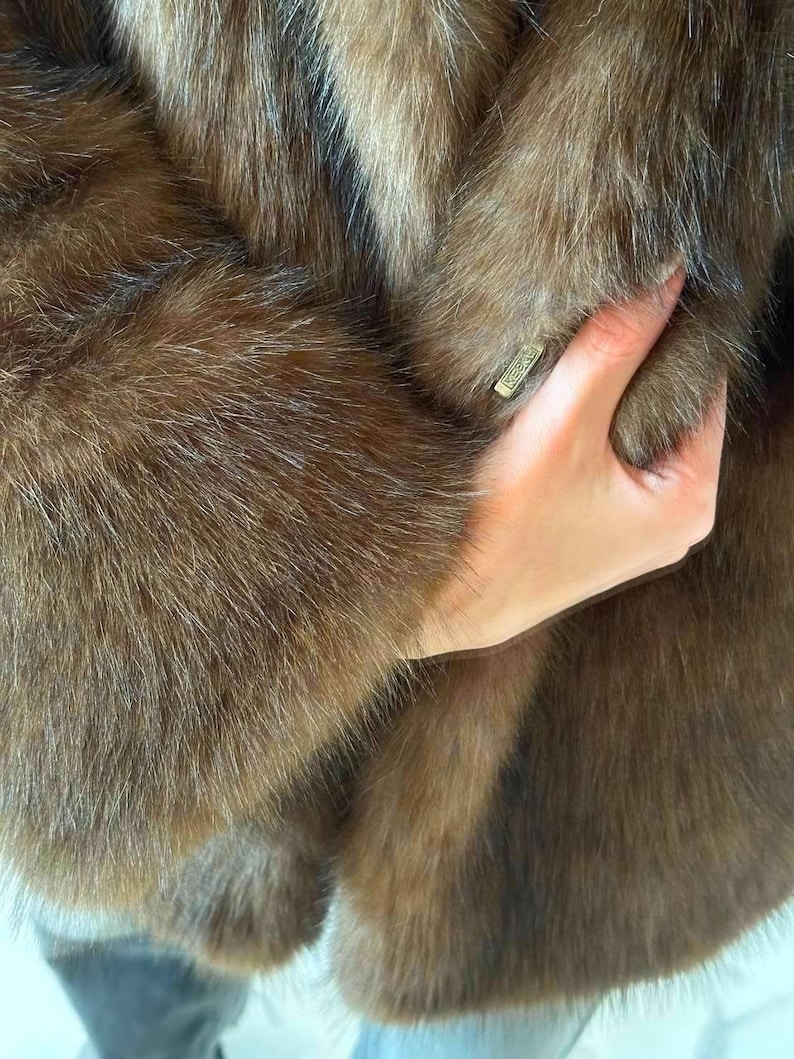 May include: Close-up of a brown fur coat. The coat has a soft, textured appearance with varying shades of brown. A hand is holding the coat, and a small, rectangular tag is visible.