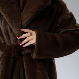 May include: A luxurious, dark brown faux fur coat with a shawl collar and a matching belt. The coat has long sleeves and a soft, plush texture. The image shows the coat's details, including the belt tied at the waist.