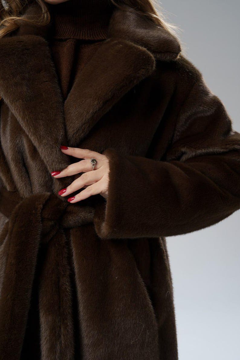 May include: A luxurious, dark brown faux fur coat with a shawl collar and a matching belt. The coat has long sleeves and a soft, plush texture. The image shows the coat's details, including the belt tied at the waist.
