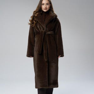 May include: A long, brown faux fur coat with a shawl collar and a matching belt. The coat is floor-length and has long sleeves. The model is wearing black pants and a brown turtleneck. The coat is a luxurious outerwear piece.