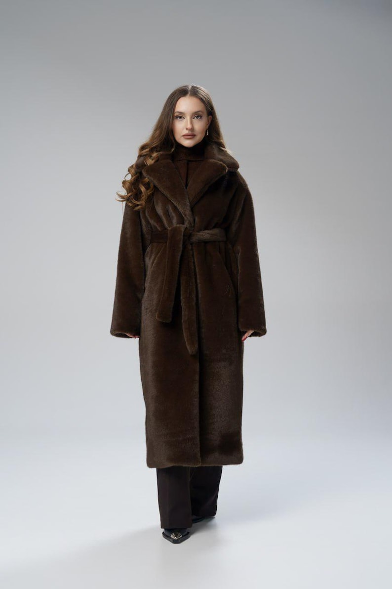 May include: A long, brown faux fur coat with a shawl collar and a matching belt. The coat is floor-length and has long sleeves. The model is wearing black pants and a brown turtleneck. The coat is a luxurious outerwear piece.
