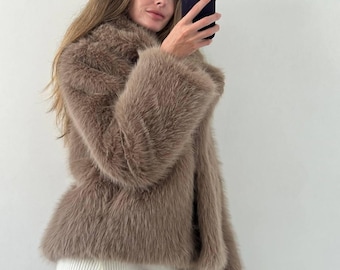 May include: A brown faux fur jacket with wide sleeves. The jacket is paired with a white ribbed knit skirt. The person is holding a black phone and taking a selfie.