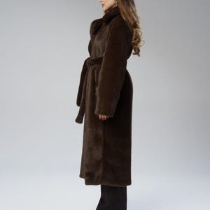 May include: A long, dark brown faux fur coat with a matching belt. The coat has a wide collar and long sleeves. The model is wearing black pants and black shoes. The coat is a stylish outerwear garment.