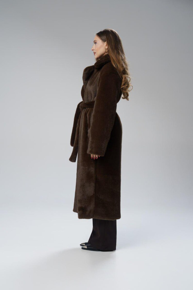 May include: A long, dark brown faux fur coat with a matching belt. The coat has a wide collar and long sleeves. The model is wearing black pants and black shoes. The coat is a stylish outerwear garment.