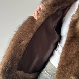 May include: A brown faux fur coat with a dark brown satin lining. The coat has a plush, textured appearance and is open, revealing the interior lining. The fur is a rich, warm brown color.