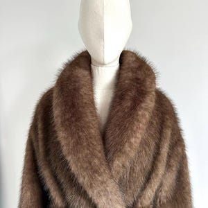 May include: A brown faux fur coat with a wide collar and a belted waist. The coat is displayed on a mannequin against a white background. The coat's fur has a soft, textured appearance.