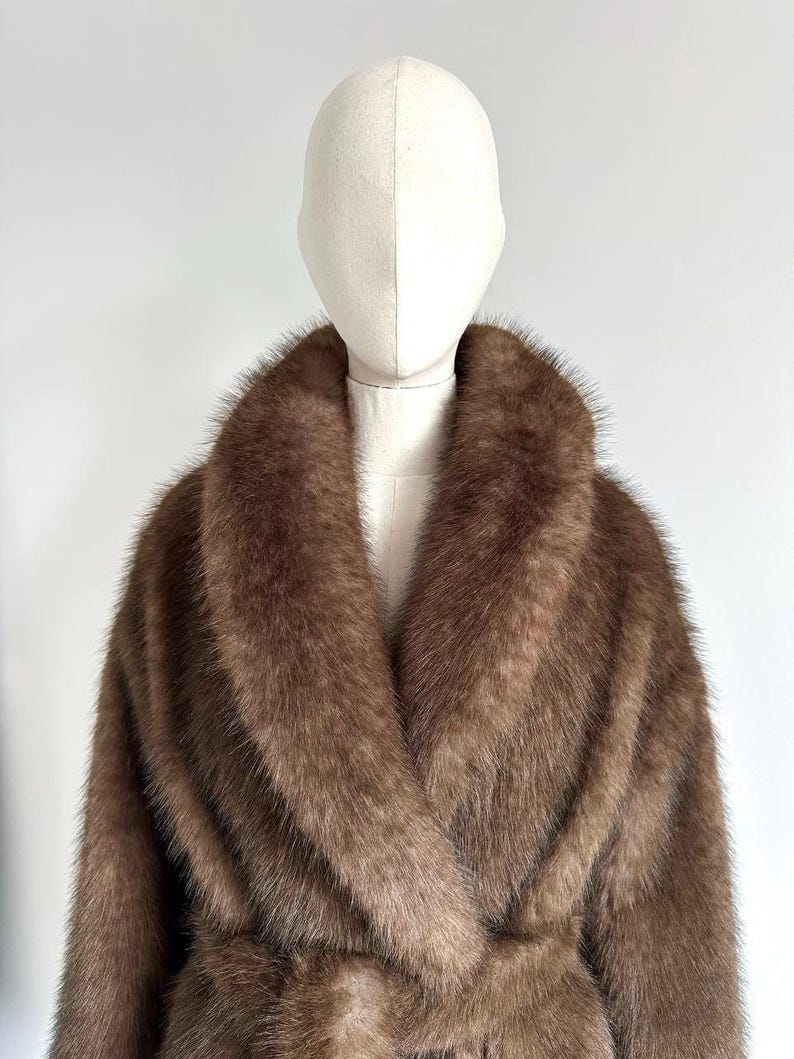 May include: A brown faux fur coat with a wide collar and a belted waist. The coat is displayed on a mannequin against a white background. The coat's fur has a soft, textured appearance.
