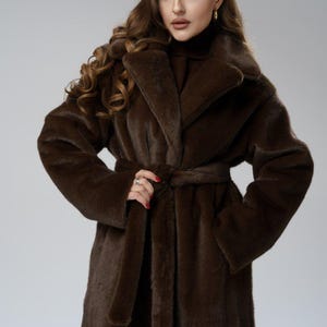 May include: A long, brown faux fur coat with a wide collar and a matching belt. The coat is knee-length and has long sleeves. The model is wearing a turtleneck sweater underneath. The coat is a rich, dark brown color.
