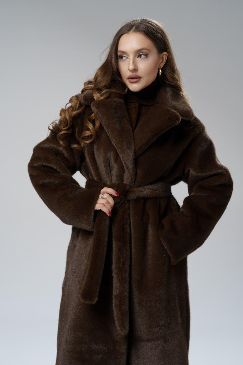May include: A long, brown faux fur coat with a wide collar and a matching belt. The coat is knee-length and has long sleeves. The model is wearing a turtleneck sweater underneath. The coat is a rich, dark brown color.