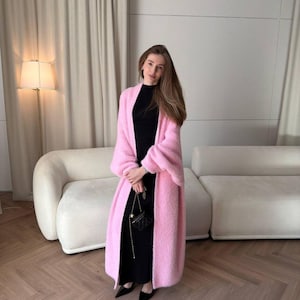 May include: A woman in a long, pale pink, fluffy cardigan over a black dress, holding a small black purse with a gold chain. She wears black pointed-toe shoes. The background features a white sofa and a floor lamp.