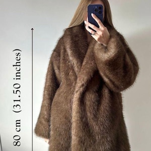 May include: A brown faux fur coat, approximately 31.50 inches in length, with a wide collar and long sleeves. The coat has a soft, textured appearance and is worn open. The overall style is a classic, oversized design.