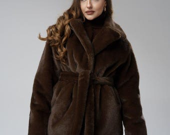 May include: A brown faux fur coat with a shawl collar and a matching belt. The coat has long sleeves and a soft, textured appearance. The model is wearing a brown turtleneck and dark pants.