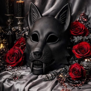 May include: A black wolf mask with sharp teeth, surrounded by red roses and jewelry, rests on a silver satin surface. Two lit black candles and a decorative bottle are in the background, creating a gothic aesthetic.