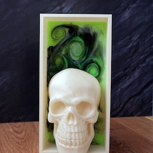 Könnte beinhalten: Eine weiß-perlmuttfarbene Totenkopfskulptur in einem weißen rechteckigen Rahmen. Der Hintergrund zeigt ein wirbelndes grün-schwarzes Design. Die Skulptur steht auf einer Holzoberfläche.