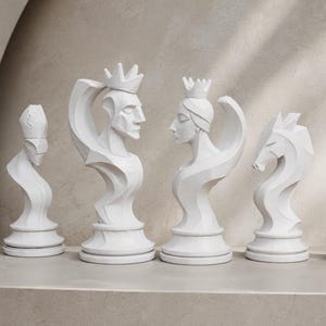 Op de afbeelding: Een set van zes witte schaakstukken met gestileerde, abstracte bustes van een pion, loper, koning, dame, paard en toren. Elk stuk heeft een gladde, matte afwerking en geometrische, vloeiende lijnen die de vorm van elk figuur bepalen.