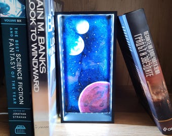 May include: A rectangular light box featuring a deep blue space scene with three planets and small white stars. The box is framed in black and illuminated from within by LED lights, positioned between two science fiction books.