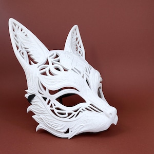 Peut inclure: Un masque de renard blanc présentant des motifs découpés complexes et des détails texturés rappelant la fourrure. Le masque possède de grandes oreilles pointues, des ouvertures oculaires en amande et une sangle élastique noire sur le côté.
