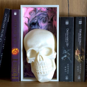 May include: A white skull sculpture inside a white rectangular frame with a pink, black, and purple swirled background. Books with titles like "Of Metal and Magic", "Hobbit", "Return of the King", and "Two Towers" are arranged on a wooden shelf.