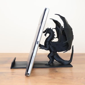 May include: A black dragon-shaped tablet stand. The dragon has large wings and is holding a tablet in its claws. The stand is made of black plastic and is designed to hold a tablet upright.