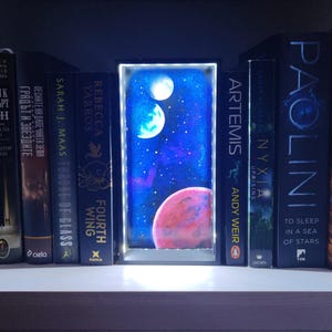 May include: A bookshelf with several books and a lighted space-themed art piece. The art piece depicts a night sky with planets and stars. The books have titles like "Artemis" and "Throne of Glass".
