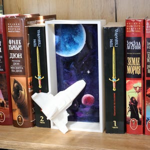 May include: A white 3D-printed spaceship model mounted on a rectangular white frame containing a deep space scene with a large blue planet, a smaller red planet, and stars, positioned between several hardcover books on a wooden shelf.
