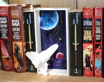 May include: A white 3D-printed spaceship model mounted on a rectangular white frame containing a deep space scene with a large blue planet, a smaller red planet, and stars, positioned between several hardcover books on a wooden shelf.